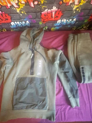 Nike Air Max grön loungewear set - Nike Air Max loungewear set i militärgrönt med matchande hoodie och byxor. Hoodien har halv dragkedja, stor magficka och tryckt Air Max-logga på bröstet. Byxorna har resår i midjan och muddar vid benslut. Perfekt för chill dagar hemma.