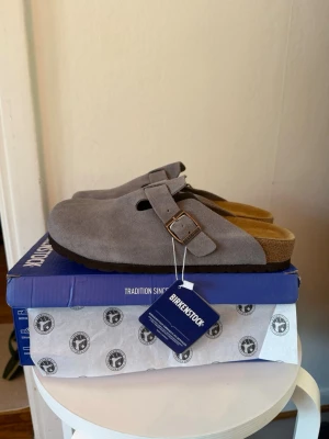 Birkenstock Boston mörkgrå tofflor - Säljer ett par Birkenstock Boston tofflor i mörkgrå mocka med justerbart spänne i metall. Klassisk slip-in modell med bekväm korksula och mjuk innersula. Perfekta för dig som gillar stilrena och tidlösa skor.