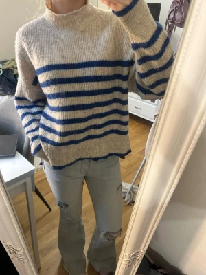Beige stickad tröja med blå ränder - Stickad tröja i beige med blå horisontella ränder. Tröjan har en lös passform, hög hals och slitsar i sidorna. 
