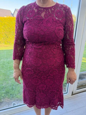 Vinröd spetsklänning med lång ärm - Vinröd klänning i spets med blommigt mönster och lång ärm. Klänningen är knälång och har en figurnära passform. Baksidan har en liten öppning med knapp i nacken och dragkedja. Perfekt för dig som vill sticka ut med elegans.