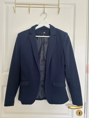 Mörkblå kavaj från H&M - Stilren mörkblå kavaj från H&M med klassisk passform. Kavajen har ett knappfäste framtill, två fickor med lock och randigt innerfoder. Perfekt för en snygg och uppklädd look. Materialet är mjukt och följsamt.