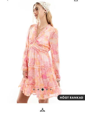 Blommig kortklänning med volang - Helt slutsåld! Supersöt kortklänning med blommigt mönster i rosa, orange och vit. Klänningen har v-ringning, volangdetaljer och långa ärmar i transparent material. Perfekt för sommarens alla tillfällen!