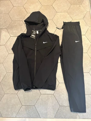  Nike Miler Svart - Snyggt svart träningsset från Nike med jacka och byxor. Jackan har huva, dragkedja och Nike-logga på bröstet. Byxorna har resår i midjan, dragsko och vit Nike-logga på låret. Perfekt för träning eller chill. Materialet är lätt och andas.