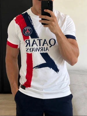 PSG t-shirt med tryck och logga - Vit PSG t-shirt med stora tryck i rött och marinblått framtill, samt klubbens logga på bröstet. T-shirten har korta ärmar med kontrastfärgade muddar och är tillverkad i mjuk bomull. Perfekt för dig som vill visa ditt stöd för laget.