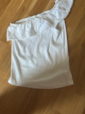 Vit offshoulder topp med spetsvolang - Snygg vit offshoulder topp med bred spetsvolang upptill. Ribbad struktur i tyget och ärmlös design som ger en fräsch och somrig känsla. Perfekt att matcha med jeans eller kjol för en trendig look.
