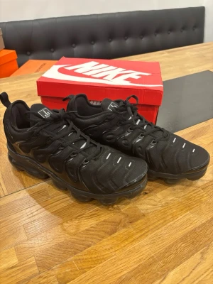 Nike Air VaporMax Plus  - Svarta Nike Air VaporMax Plus sneakers med futuristisk design, synliga luftbubblor i sulan och detaljer i svart och grått. Ovandel i textil och syntet med reflekterande inslag och snörning. Perfekta för dig som gillar streetwear och sportig stil.