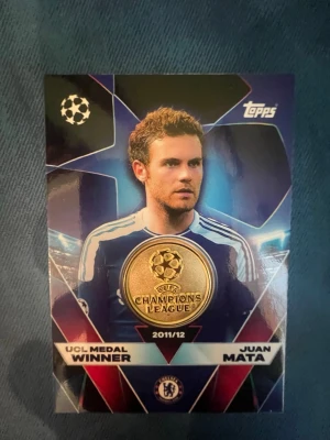 UCL Medal Winner kort Juan Mata - Samlarobjekt: UCL Medal Winner-kort från Topps Match Attax med Juan Mata, Chelsea, säsongen 2011/12. Kortet har en guldig Champions League-medalj på framsidan och är gjort av glansig kartong i blå, guld och vita färger. Perfekt för fotbollsfans och samlare.
