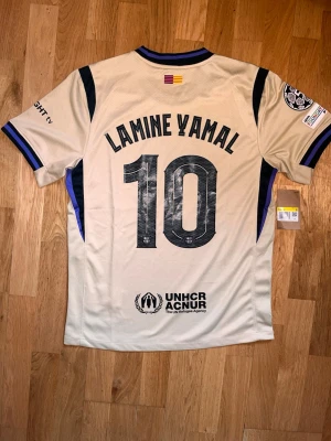 FC Barcelona Lamine Yamal tröja - Snygg beige FC Barcelona matchtröja med Lamine Yamal och nummer 10 på ryggen. Tröjan har svarta och lila detaljer, klubbmärke och sponsorloggor. Korta ärmar och rund hals. Perfekt för fotbollsfans som vill sticka ut på planen eller läktaren. Aldrig använt 10/10 skicka, priset går att diskuteras!