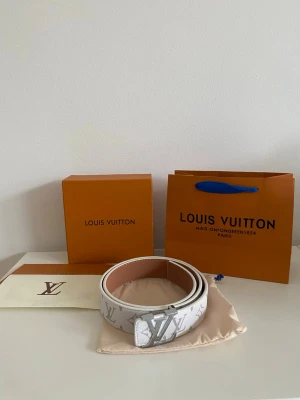vitt skärp med monogram - Stilrent vitt skärp  med klassiskt monogram i silver på spännet. Skärpet är gjort i exklusivt skinn och har en beige insida. Perfekt accessoar för att lyfta din outfit med en lyxig touch.