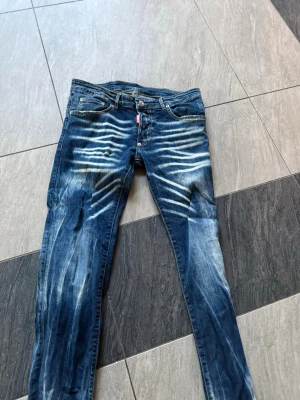 Blå Dsquared2 jeans med slitningar - Säljer ett par blå jeans från Dsquared2 med coola slitningar och tvättade detaljer. Jeansen har smal passform, klassisk femficksmodell och knappgylf. Materialet är denim i bomull och de har en röd Dsquared2-logga vid gylfen.