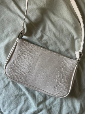 Beige axelväska med krokopräglat mönster - Snygg beigevit axelväska i fejkläder med krokopräglat mönster. Väskan har en enkel, stilren design med dragkedja upptill axelrem.