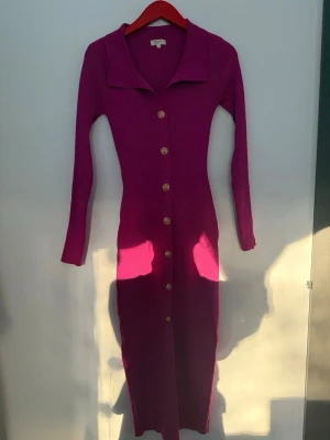 Långärmad rosa ribbad klänning - Snygg långärmad klänning i stark rosa färg med ribbad struktur. Klänningen har en klassisk krage och knappar hela vägen framtill. Figurnära modell som ger en trendig siluett. Perfekt för dig som gillar färg och vill sticka ut.