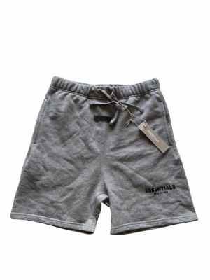 Essentials FOG short - Snygga grå shorts från Essentials Fear of God med snörning i midjan och logga tryckt på benet. Mjukt material och avslappnad passform, perfekta för chill dagar. Elastisk midja och sidofickor för extra komfort. Har aldrig använts
