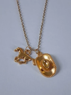 Guldigt halsband med häst och cowboyhatt - Unikt halsband i guldfärgad rostfritt metall med två hängen: en häst och en detaljerad cowboyhatt. Kedjan är tunn och smidig, och motiven ger en cool westernkänsla. Perfekt för dig som gillar hästar eller vill ha en personlig accessoar med westernstil.