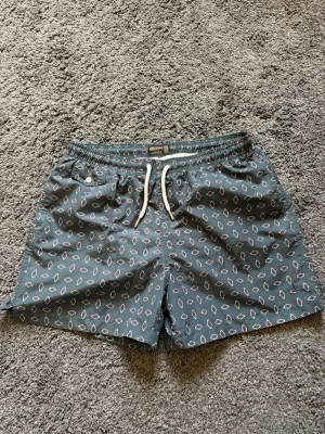 Mörkgröna badshorts med mönster från Morris - Snygga mörkgröna badshorts från Morris med vitt geometriskt mönster. Shortsen har resår och dragsko i midjan samt sidofickor och en bakficka. Perfekta för stranden eller poolen, tillverkade i snabbtorkande material. Nypris 1199kr.