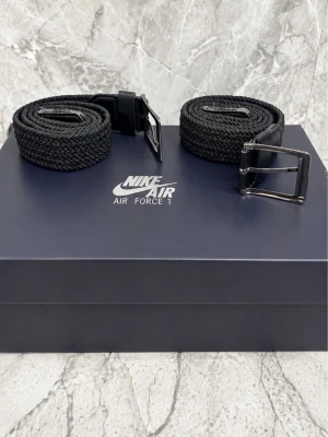 Svarta flätade bälten från Nike - Två svarta flätade bälten från Nike med klassiskt spänne i metall och detaljer i skinn. Perfekta accessoarer för att lyfta din outfit med en sportig touch. Snygg och enkel design som passar till det mesta.90-100cm