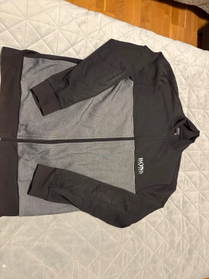 Svart och grå zip-hoodie från BOSS - Snygg zip-hoodie från BOSS i svart och grått med vit logga på bröstet. Tröjan har lång ärm, ribbade muddar och dragkedja hela vägen. Perfekt för en stilren och avslappnad look.