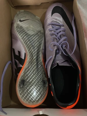 Nike Mercurial lila fotbollsskor - Snygga Nike Mercurial fotbollsskor i lila med svarta detaljer och orange inslag vid hälen. Skorna har en modern, aerodynamisk design med genomskinlig sula och klassisk snörning. Perfekta för dig som vill sticka ut på planen och få grymt grepp på underlaget.
