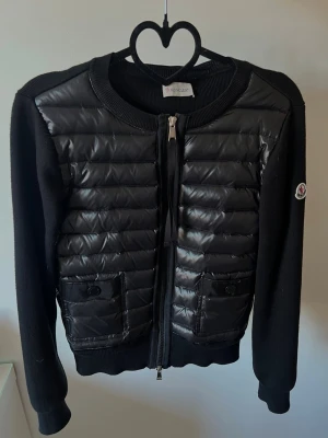 Svart cardiganjacka från Moncler - Snygg svart cardiganjacka från Moncler med quiltad framsida i blankt material och stickade ärmar. Jackan har två fickor framtill med knappstängning och dragkedja hela vägen. 