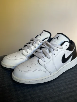 Air Jordan 1 Low svart/vit sneakers - Snygga Air Jordan 1 Low sneakers i svart och vitt läder med klassisk Swoosh-logga på sidan och Air Jordan Wings-logga bak. Skorna har perforerad tå, vita skosnören och svart yttersula. Perfekta för dig som gillar streetwear och ikonisk design. Knappt använda, har ej kvar lådan
