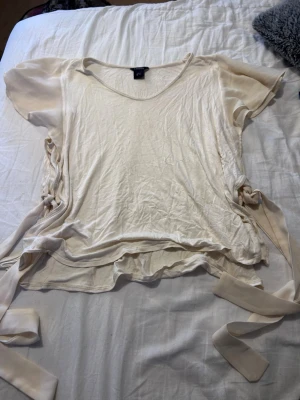 Krämvit topp med knyt från Lindex - Krämvit topp från Lindex med korta ärmar och v-ringning. Snygga knytband i sidorna ger en unik look. Tunt och mjukt material, perfekt för varmare dagar. Enkel att styla till olika outfits.