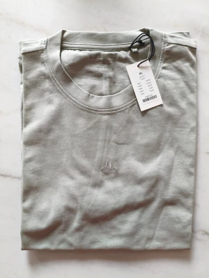 Ljusgrå t-shirt från New Garments - Säljer en stilren ljusgrå t-shirt från New Garments i mjuk bomull. T-shirten har rund halsringning och ett diskret broderat smiley-ansikte på bröstet. Enkel och clean design som passar till allt.