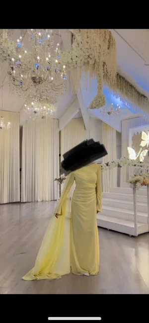 Ljusgul långklänning med släp - Elegant ljusgul långklänning med ett axelbandslöst snitt på ena sidan och lång ärm på andra. Klänningen har ett dramatiskt släp och är figurnära med draperade detaljer. Perfekt för speciella tillfällen. Passar S och M