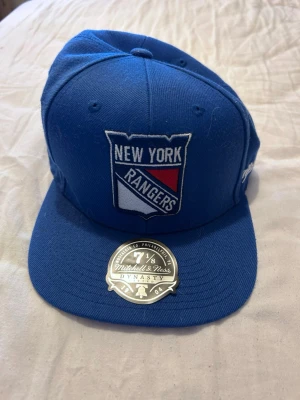 Blå New York Rangers keps - Snygg blå keps från Mitchell & Ness med New York Rangers-logga framtill och jubileumsmärke på sidan. Klassisk flat brim och broderade detaljer. Perfekt för dig som gillar hockey och streetwear.