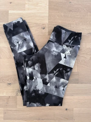 Träningstights från Adidas - Snygga träningstights från Adidas med svart, grått och vitt grafiskt mönster. Hög midja och stretchigt material. Adidas-logga tryckt på låret. Endast använda 2-3 gånger och är i väldigt fint skick.