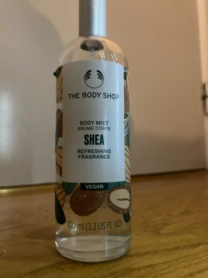 The Body Shop Shea Body Mist 100 ml - Fräsch body mist från The Body Shop med doft av shea. Kommer i en genomskinlig glasflaska med silverfärgad spraykork och dekorativa illustrationer. Innehåller 100 ml och är vegansk med 97% ingredienser av naturligt ursprung.