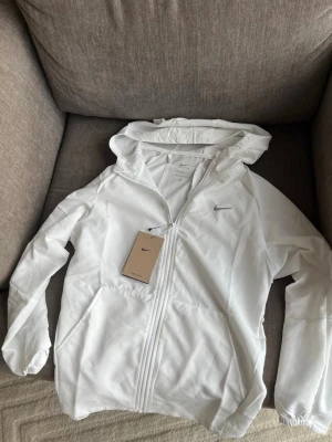 Vit vindjacka från Nike Dri-FIT - Snygg vit vindjacka från Nike med huva och dragkedja framtill. Tillverkad i lätt och funktionell polyester med Dri-FIT-teknologi som håller dig torr. Jackan har diskret Nike-logga på bröstet och praktiska fickor på sidorna.