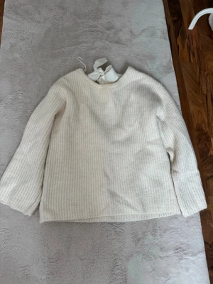 Beige stickad tröja med rosett - Säljer en beige stickad tröja med ribbad struktur och rund hals. Tröjan har en snygg öppen rygg med en stor rosett i nacken som detalj. Långa ärmar och rak passform gör den både stilren och trendig. Perfekt för dig som gillar en enkel men söt look.