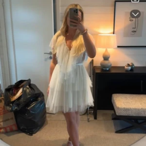 Vit kortklänning från Ida Sjöstedt - Superfin vit kortklänning från Ida Sjöstedt med lager på lager av tunn tyll, v-ringning och volangdetaljer. Klänningen har korta ärmar och ett knytband i midjan som ger en romantisk och luftig känsla. Perfekt för dig som vill sticka ut med en drömmig look. Aldrig använd. Jag är 171 cm lång och bär storlek 40.