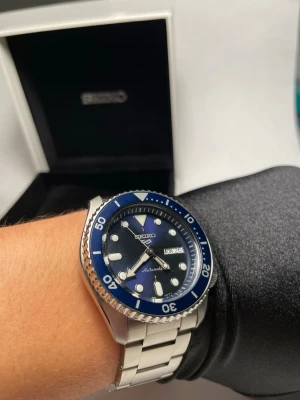 Seiko automatisk klocka blå/silver - Full set - Snygg automatisk klocka från Seiko med blå urtavla och matchande blå vridring. Klockan har tydliga indexmarkeringar, dag- och datumvisning samt robust länkarmband i silverfärgat stål. Perfekt för dig som gillar klassisk men sportig stil.