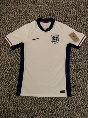 England Nike fotbollströja vit/blå - Snygg England fotbollströja från Nike i vitt med marinblå detaljer längs sidorna och ärmarna. Tröjan har korta ärmar, broderad Nike-logga och Englands landslagsemblem på bröstet. Materialet är lätt och andas, perfekt för träning eller match.