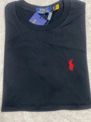 Svart t-shirt från Polo Ralph Lauren - Klassisk svart t-shirt från Polo Ralph Lauren med röd broderad logga på bröstet. T-shirten har rund halsringning och är tillverkad i mjuk bomull för en skön känsla. Perfekt basplagg med stilren design.