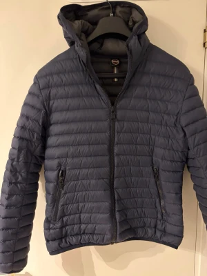 Mörkblå dunjacka från Colmar - Snygg mörkblå dunjacka från Colmar med huva och två dragkedjeförsedda fickor. Jackan har quiltad design med horisontella sömmar och Colmar-logga på ärmen. Perfekt för kalla dagar och riktigt lätt att bära.