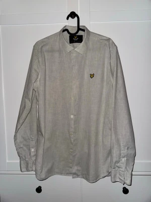 Ljusgrå skjorta från Lyle & Scott - Klassisk ljusgrå skjorta från Lyle & Scott med gul broderad logga på bröstet. Skjortan har lång ärm, knappar framtill och en stilren krage.