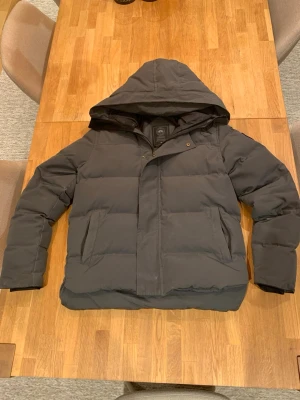 Svart pufferjacka med huva - Säljer en svart pufferjacka från canada goose  med stor huva och dragkedja framtill. Jackan har två stora fickor på sidorna och breda quiltade paneler. Perfekt för kalla vinterdagar och riktigt skön att bära. Det är en liten skada på sidan men den syns knappt inte har lagat den. Om ni vill ha fler bilder så kan ni fråga 