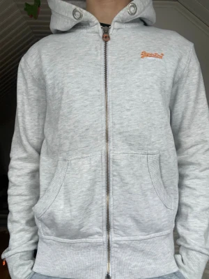 Ljusgrå zip-hoodie från Superdry - Ljusgrå hoodie med dragkedja från Superdry Orange Label. Klassisk modell med huva, två stora fickor fram och diskret orange Superdry-logga på bröstet. Tillverkad i mjuk bomullsblandning för skön känsla. Perfekt för en avslappnad streetwear-look.