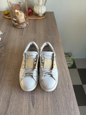 Valentino vita sneakers med beige detalj - Säljer ett par vita sneakers från Valentino med bred beige rem över ovandelen och silverfärgade nitar på sidan. Skorna har klassisk låg profil, rund tå och vita snören. Tillverkade i läder för en lyxig känsla och stilren look.