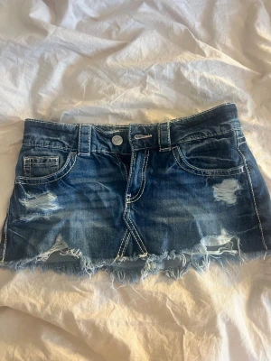 Blå jeanskjol med slitningar - Säljer en kort blå jeanskjol med slitna detaljer och fransig nederkant. Kjolen har bakfickor med strass och mönster. En perfekt kjol för dig som gillar minikjolar!