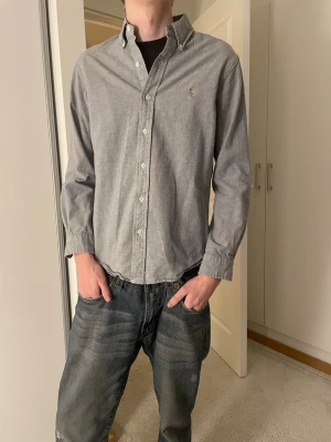 Grå skjorta från Ralph Lauren - Säljer en sjukt snygg grå skjorta från Ralph Lauren med klassisk button-down krage och broderad logga på bröstet. Skjortan är i bomull och har slim fit passform samt vita knappar framtill. Perfekt för en clean och enkel look.