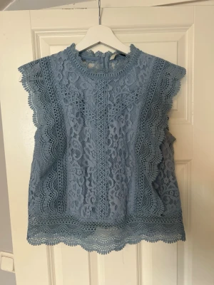 Blå spetsblus med volangdetaljer - Supersöt blå spetsblus med volangkant med medelhög hals. Blusen är ärmlös och har fina spetsmönster över hela plagget samt knyt i nacken. Perfekt för dig som vill piffa upp din outfit lite extra!  Använd enstaka gånger, nypris 599kr!