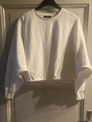 Vit strukturerad tröja från Bershka - Säljer en vit, croppad tröja från Bershka med snygg struktur och ribbade muddar. Tröjan har rund halsringning och ett diskret mönster i tyget som ger en modern vibe. Perfekt till jeans eller kjol för en clean look.