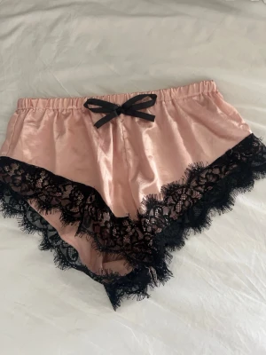 Rosa satinshorts med svart spets - Snygga loungewear-shorts i glansigt rosa satin med bred svart spetskant och en liten svart rosett framtill i midjan. Perfekta för en chill dag hemma eller mysiga kvällar. Shortsen har elastisk midja för extra komfort och en feminin look. Ingen lapp och köpt på loppis