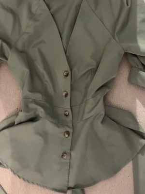 Olivgrön blus med knyt och knappar - Snygg olivgrön blus med v-ringning, knappar framtill och ett knyt i midjan. Blusen har trekvartsärmar och lite vintage vibe.