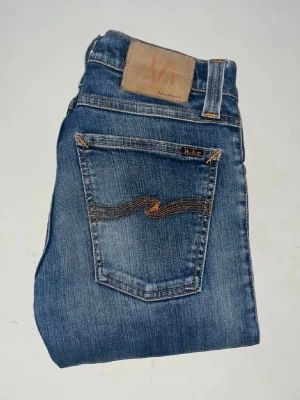 Nudie jeans  - Säljer ett par klassiska blå skinny jeans från Nudie Jeans, i modellen tajht long john. Jeansen har snygga slitningar och kontrastsömmar. Midja 33cm, längd 106cm. JAG SKICKAR EJ FLER BILDER NÄR JAG BÄR JEANSEN! för bättre inblick i passformen rekommenderar jag att googla på modellnamnet. För storleksguide kolla måtten noggrant då jeansen alltid kan vara uppsydda heller krympta och jag ej tar emot returer :) s5,2