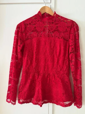 Röd spetsblus med hög krage - Snygg röd blus i spets med långärmad design och hög krage. Blusen har transparenta partier upptill och vid ärmarna, samt en figurnära passform och knapp i nacken. Perfekt för dig som vill sticka ut med en trendig och elegant look.