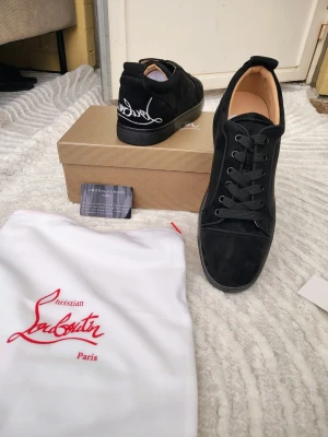 Svarta sneakers från Christian Louboutin - Snygga svarta sneakers i mocka från Christian Louboutin med svart sula och klassisk logga broderad i vitt på hälen. Skorna har svarta skosnören och en stilren, låg profil som passar till många olika outfits.